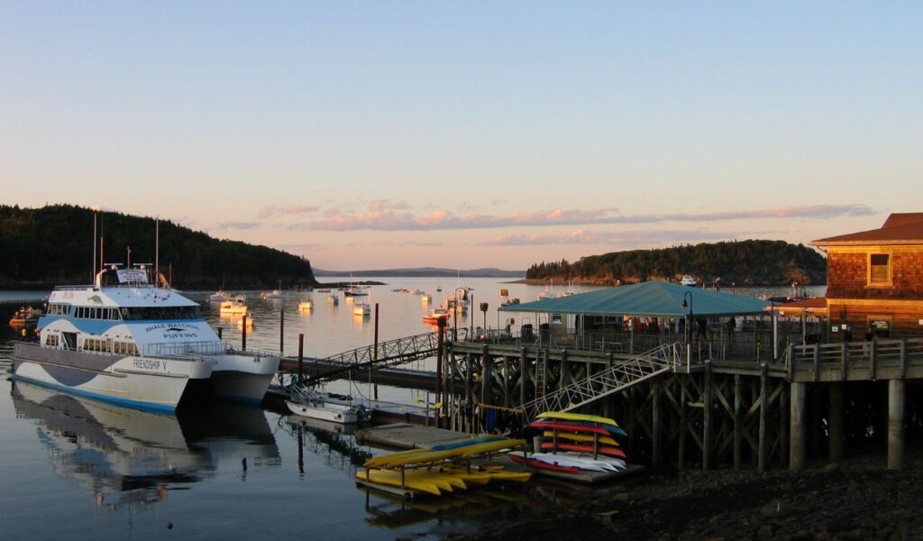 Bar Harbor waterfront at sunset with whale-watching boat, dock, kayaks, and moored boats in calm water surrounded by forested hills.