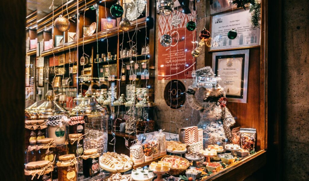 Festively decorated shop window at Bath Christmas Market, filled with jars of sweets, nuts, pastries, and hanging ornaments, with a shimmering snowman figure.