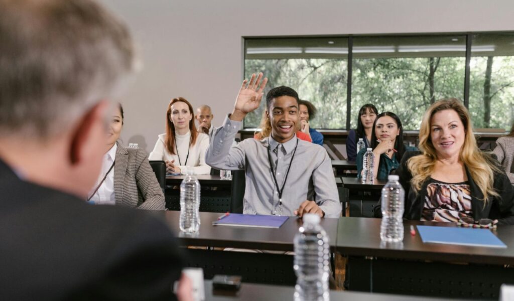 A young man raising his hand to ask a question during a seminar or conference, surrounded by other attendees seated at tables with notebooks and water bottles.