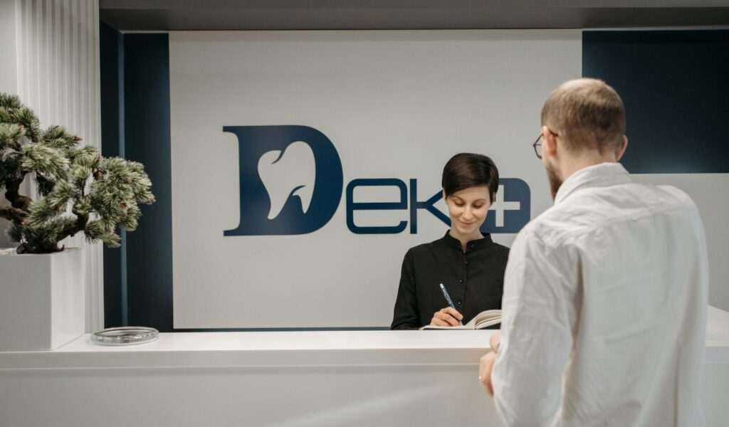 Smiling receptionist writes in a notebook while assisting a casually dressed man at a clean, modern front desk.