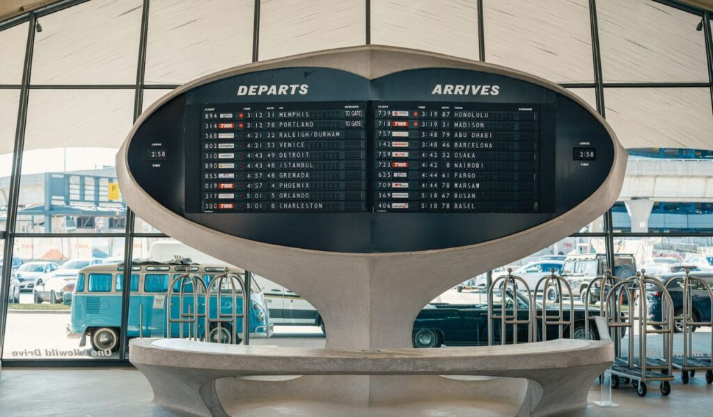 A retro-style airport departure and arrival board displays international flight times inside a terminal with large glass windows and vintage decor.