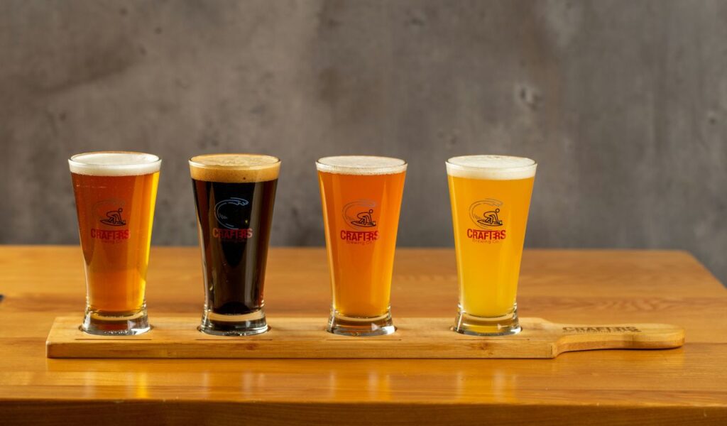 Four glasses of craft beer in different colors arranged on a wooden paddle, placed on a wooden table with a neutral gray background.