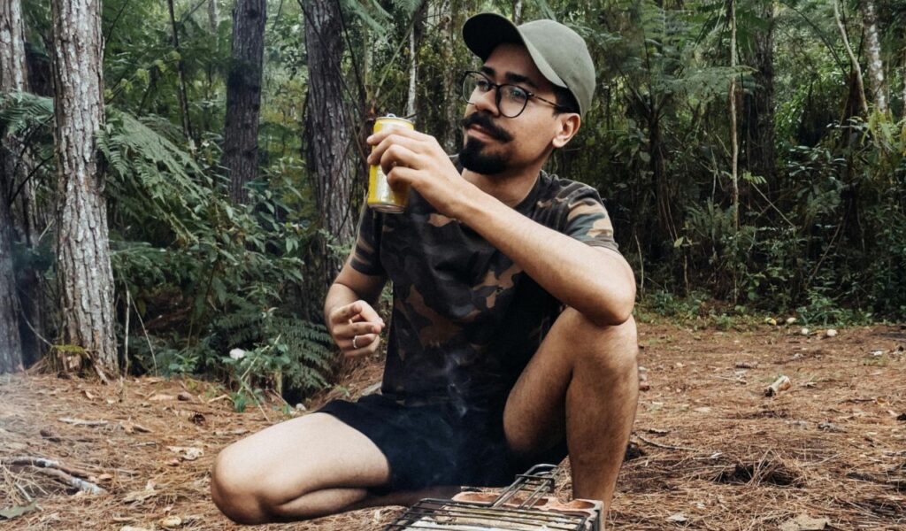 Man enjoying a beer in a forest while grilling over an open fire, capturing the spirit of beer hiking and outdoor adventure.