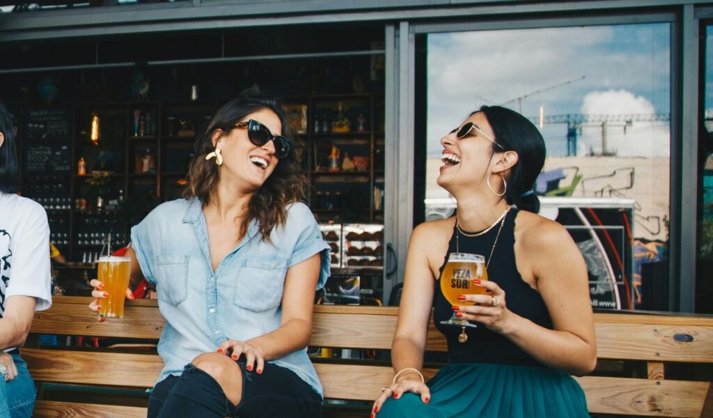 Two women sitting on a bench outside a bar, laughing and holding glasses of beer, enjoying a relaxed beer-tasting experience.
