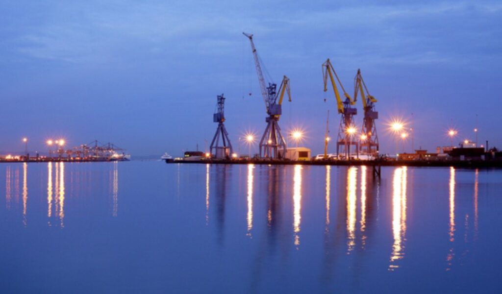 Evening view of Belfast, Maine’s harbor with industrial cranes and glowing lights reflecting off calm blue water.