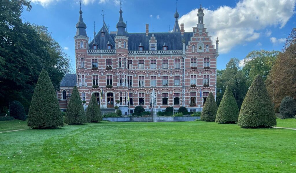A grand European château with red brick and white stone trim, pointed turrets, and manicured conical topiary trees lining a lush green lawn.