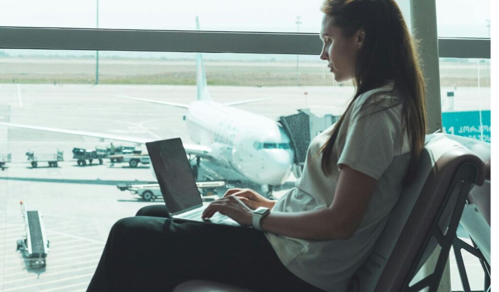 Woman sitting in airport terminal using a laptop, with a parked airplane and ground equipment visible through the window.