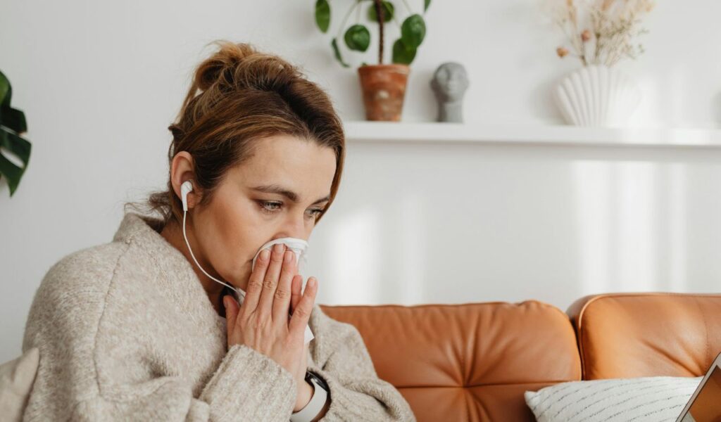 Woman sitting on a couch wearing earphones and blowing her nose into a tissue, appearing unwell or dealing with a cold, in a cozy living room setting.
