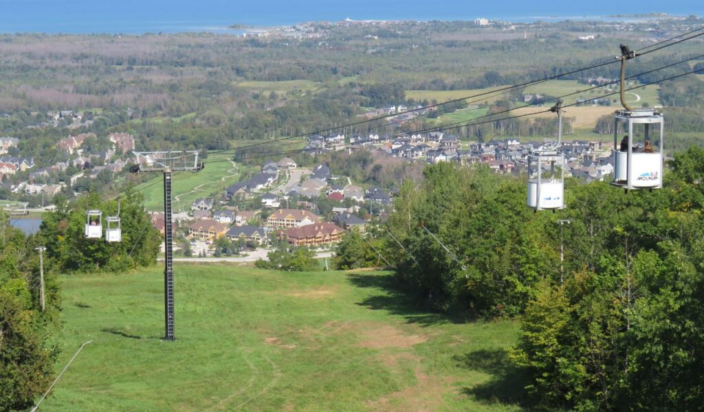  A scenic view from a gondola ride at Blue Mountain Resort, overlooking the village below with homes, trees, and Lake Huron in the distance.
