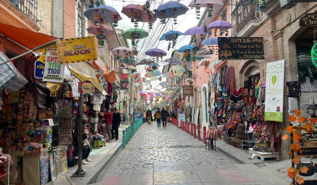 A vibrant street market in La Paz, Bolivia, with colorful umbrellas hanging overhead, shops selling local goods, and people walking along the cobblestone street