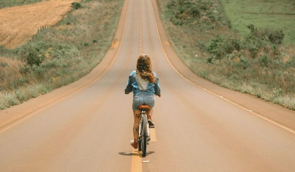 Woman cycling alone on a quiet, open country road, surrounded by fields and greenery, evoking peace and reflection.