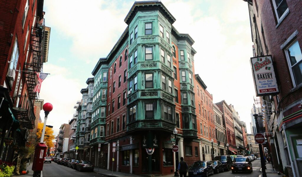 A historic street in Boston’s North End with brick buildings and green-trimmed architecture, featuring Bova’s Bakery sign, parked cars, and pedestrians under a partly cloudy sky.