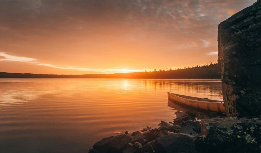 A golden canoe rests on the rocky shore of a calm lake at sunset in Minnesota's Boundary Waters Canoe Area.