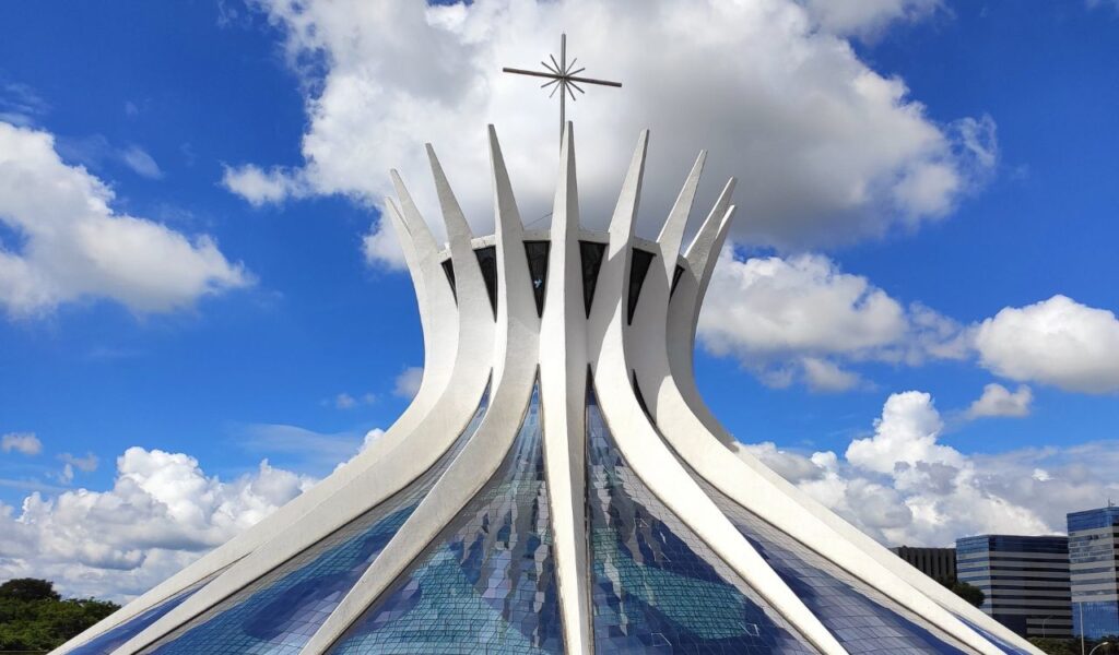 The Cathedral of Brasília, a modernist concrete and glass structure with 16 curved columns reaching skyward, topped by a metal cross against a blue sky.