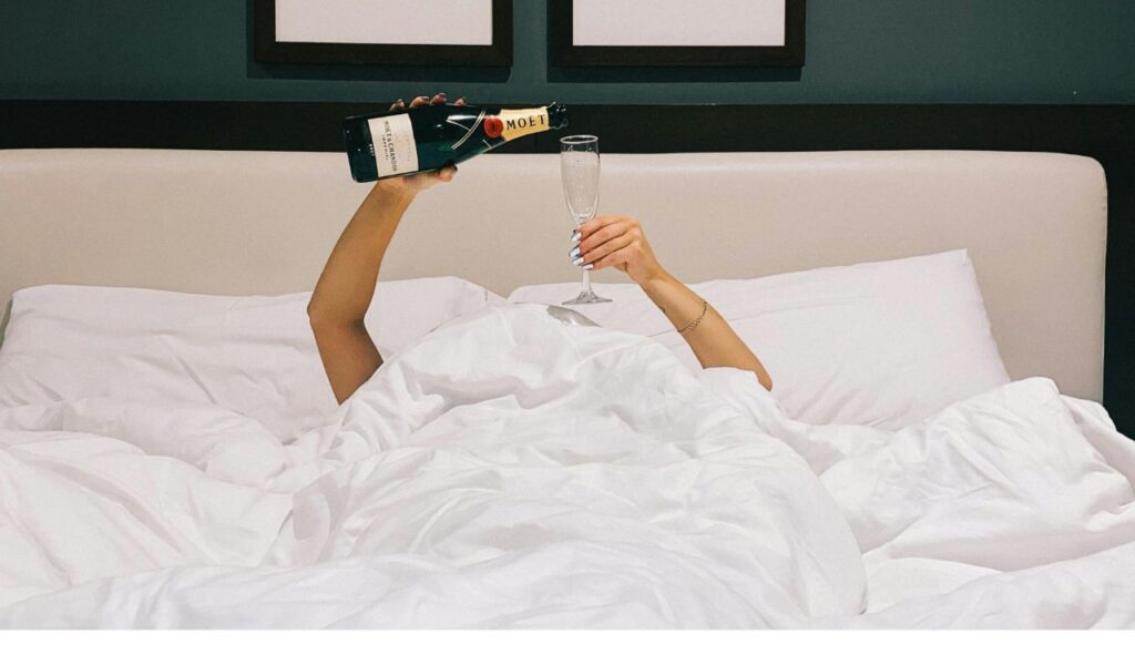  A person lying in bed under white blankets, holding a champagne bottle and flute above their head in a playful toast.