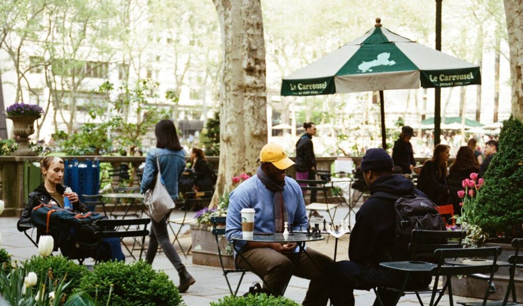 People relaxing and playing chess at outdoor tables in Bryant Park, New York City, surrounded by greenery, tulips, and a large umbrella labeled "Le Carousel.