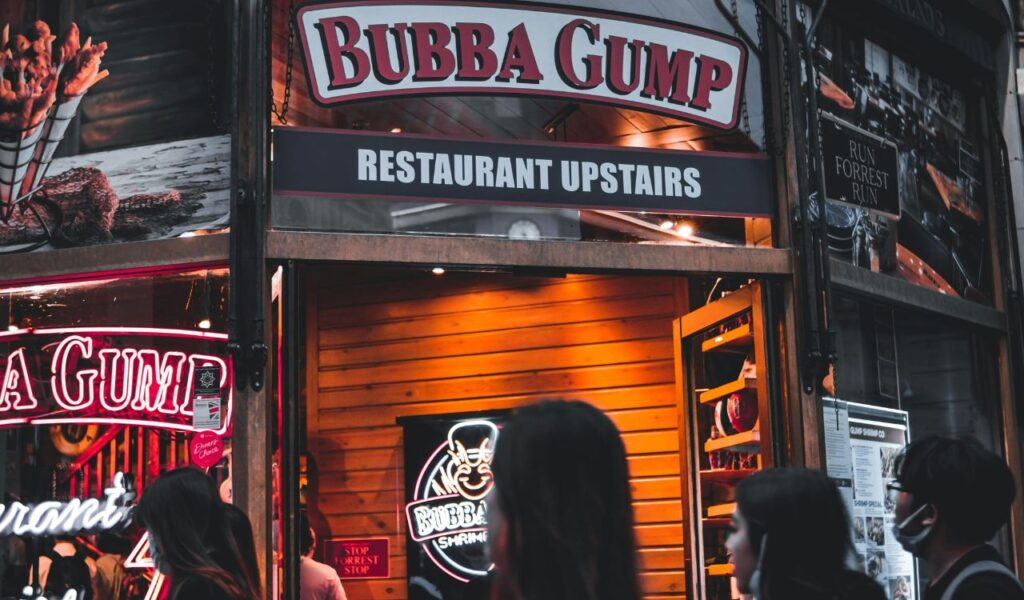 Entrance of Bubba Gump Shrimp Co. restaurant with neon signage, wooden decor, and people walking past.