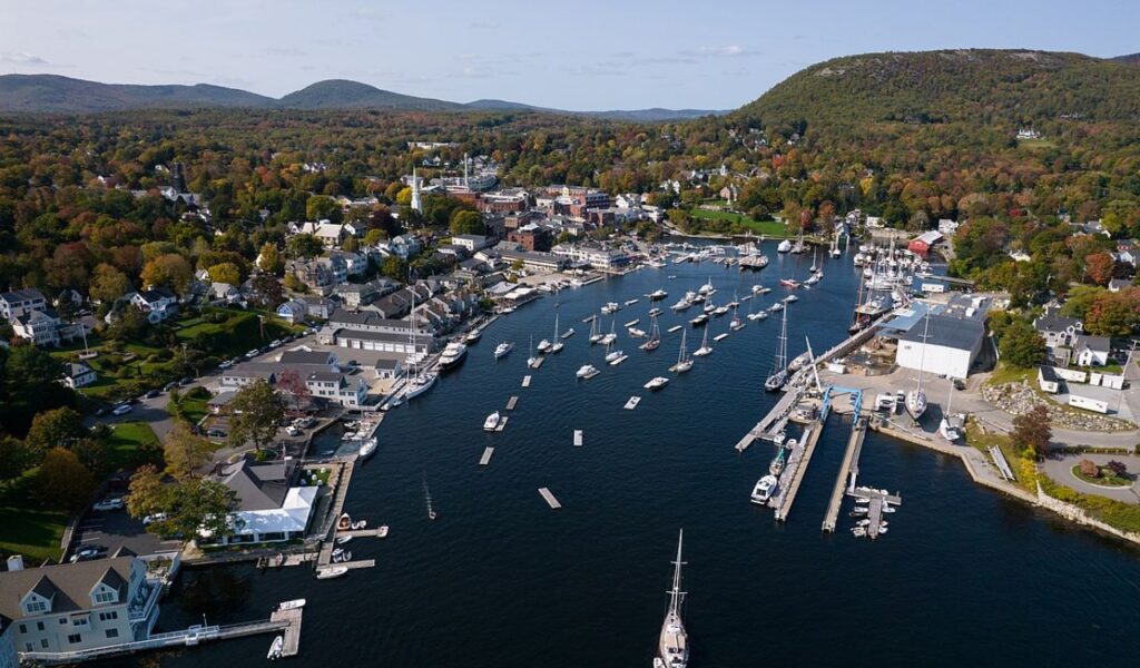 Aerial view of Camden, Maine featuring a vibrant harbor filled with sailboats, nestled among colorful fall foliage and hills.