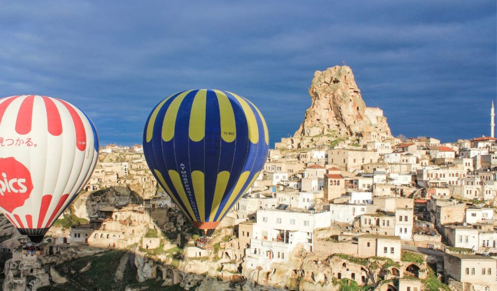 Colorful hot air balloons float over Cappadocia’s unique rock formations and stone homes under a dramatic blue sky.