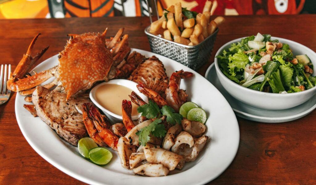 A seafood platter with grilled shrimp, fish, and crab, served with dipping sauce, lime wedges, French fries, and a green salad.