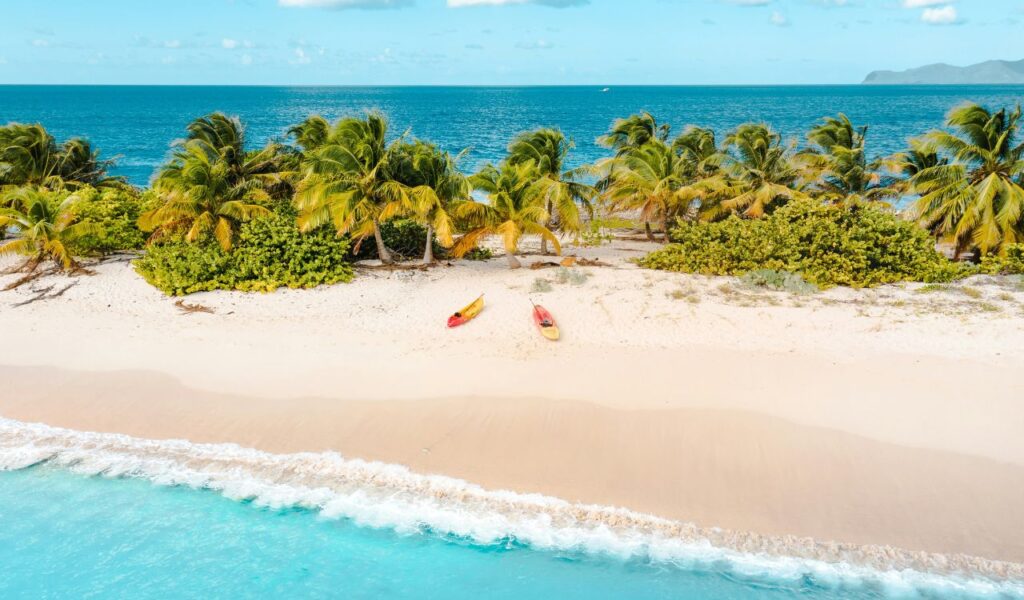 Aerial view of a pristine beach on Carriacou Island with turquoise water, white sand, palm trees, and two kayaks resting on the shore.