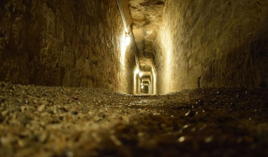 Dimly lit tunnel inside the Catacombs of Rome, with rough stone walls, arched ceiling, and gravel-covered floor.