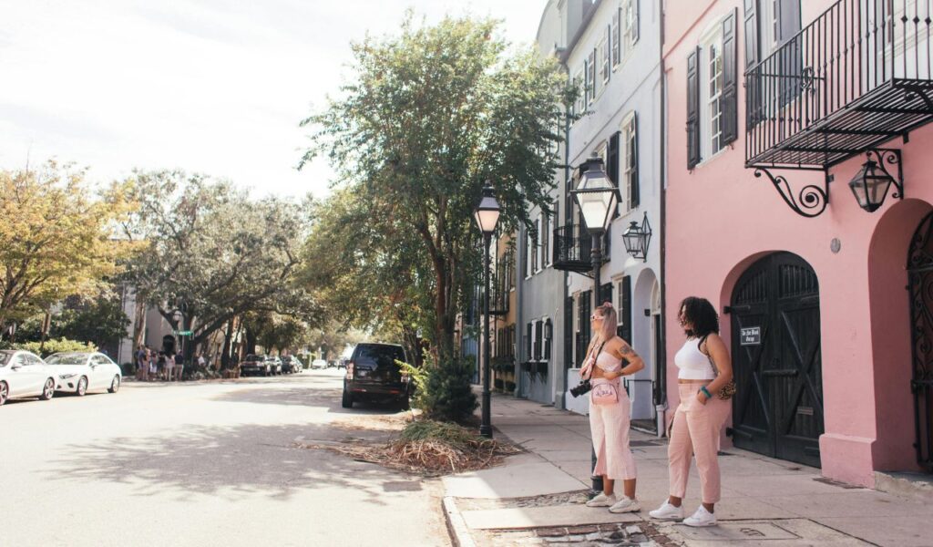 Two women stand outside pastel-colored historic homes on a sunny street in Charleston, South Carolina, near the famous Rainbow Row.