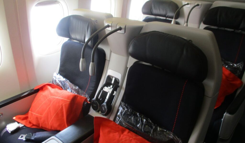 Premium airplane seats with black leather headrests, orange pillows, and individual reading lights, next to aircraft windows.