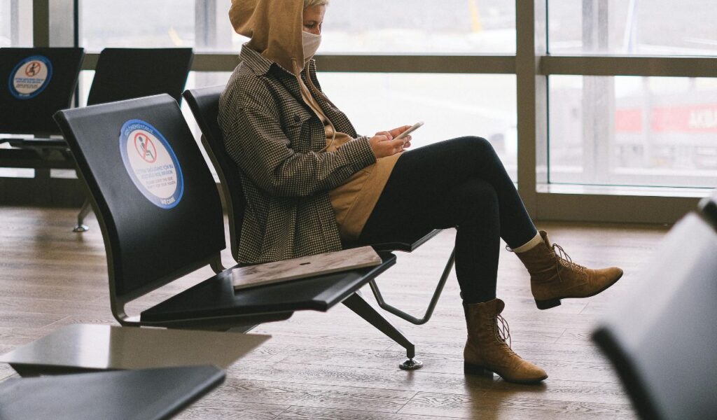 Traveler in hoodie and mask relaxes in airport lounge while checking phone, calmly waiting for flight boarding.