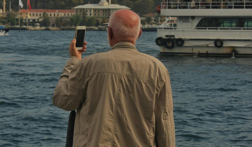 An elderly man standing by the water, holding a smartphone and taking a photo of a boat in the distance.