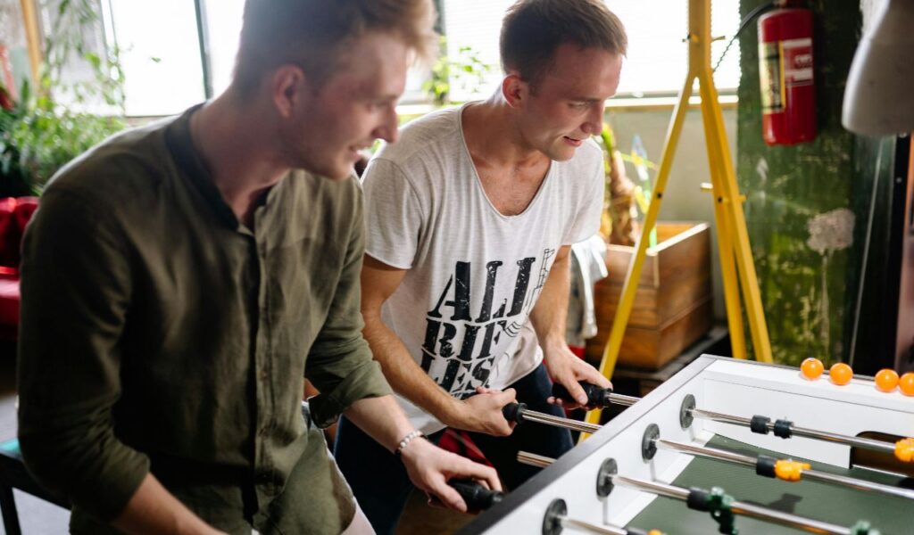 Two men smiling and playing foosball in a casual indoor setting, suggesting a relaxed environment unrelated to tourist attractions or scams.