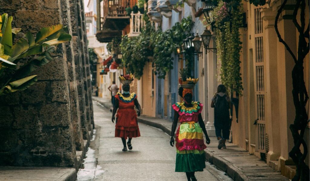 Women in colorful traditional dresses walk down a narrow street in Cartagena, Colombia, carrying fruit baskets on their heads.