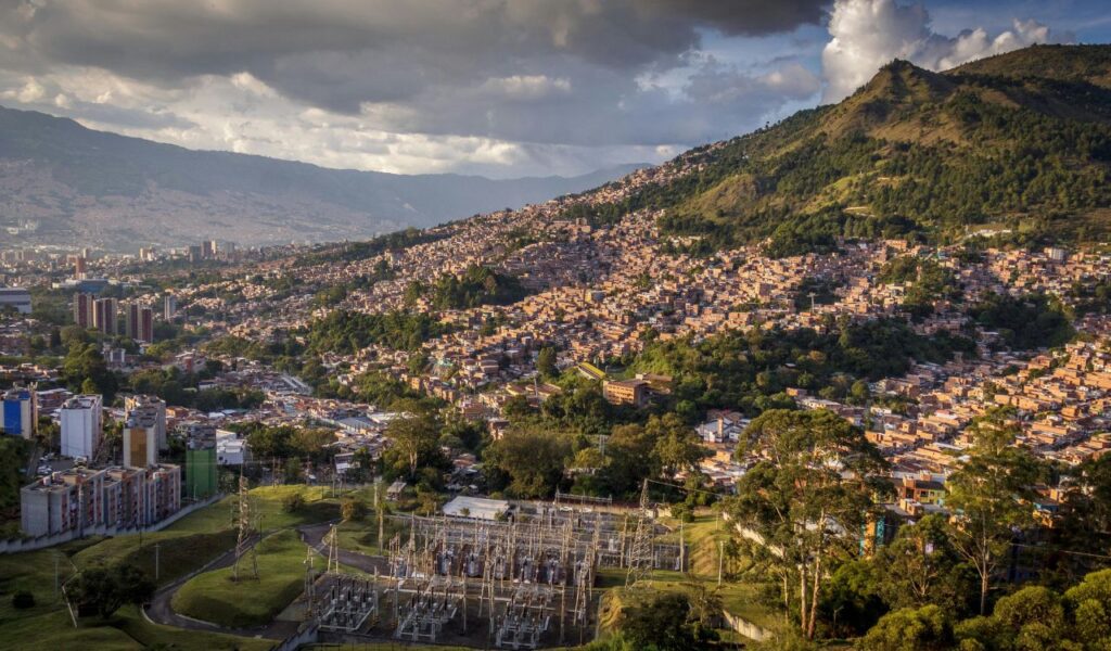 A panoramic view of Medellín, Colombia, with densely packed buildings climbing up the green hillsides, set under dramatic clouds.
