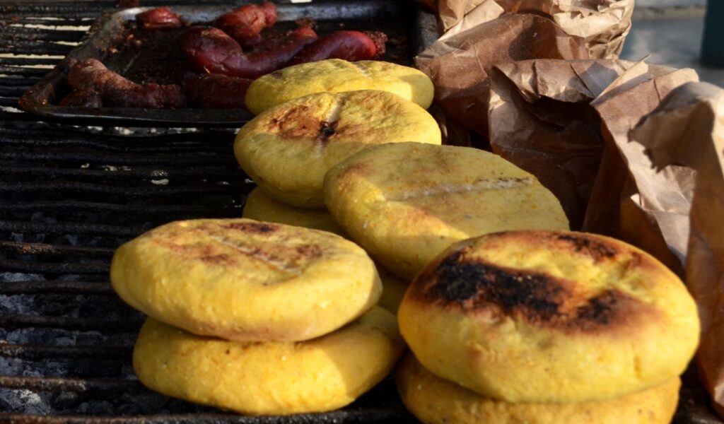 Grilled Colombian arepas stacked on a barbecue, with golden-brown crusts and sausages cooking in the background.
