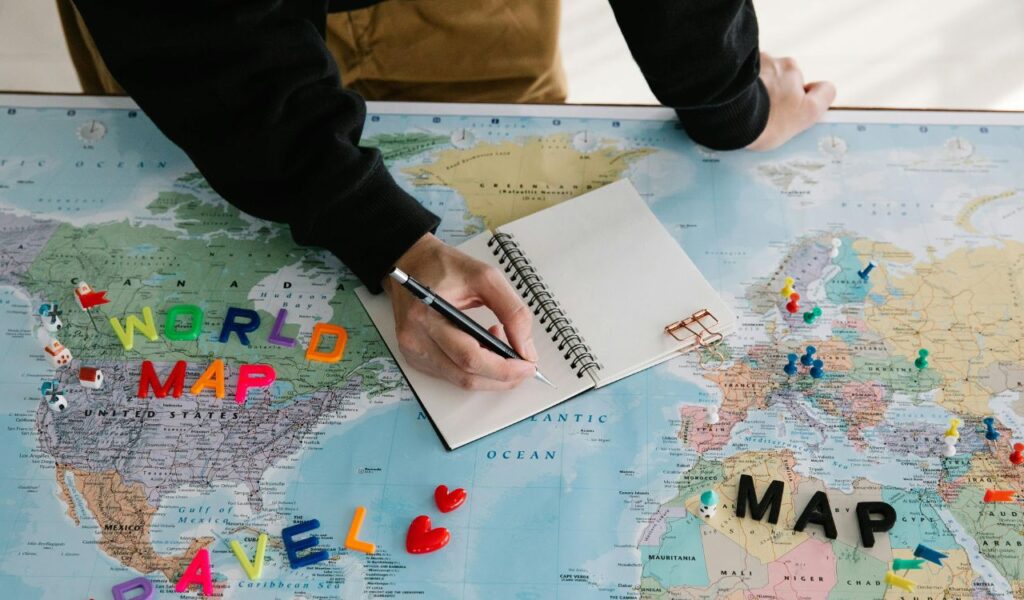 Person writing in notebook over a world map with travel pins, planning routes and preparing for a global trip.