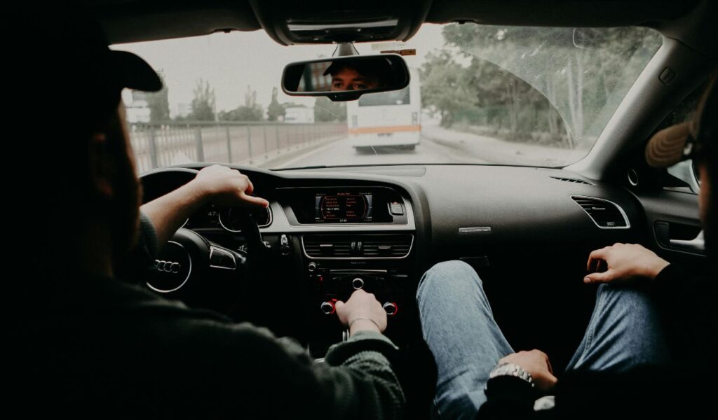 View from the backseat of a car showing driver and passenger relaxed, with dashboard and road ahead through windshield.