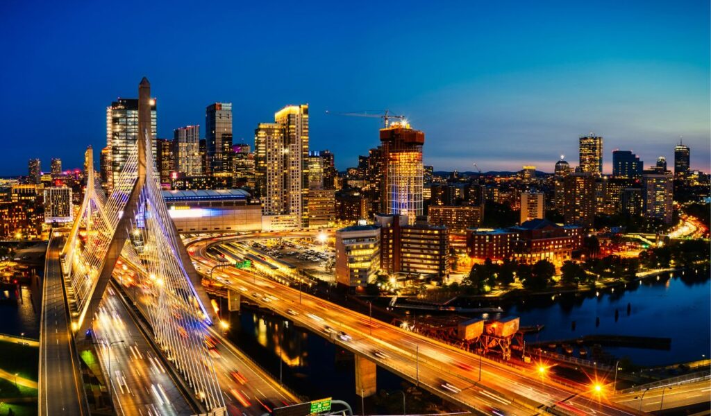 Boston skyline at night with the Zakim Bridge lit up, busy highways, glowing skyscrapers, and city lights reflecting on the river.