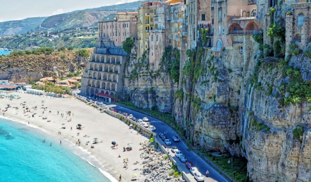 Colorful cliffside buildings overlook a sandy beach and turquoise sea, with sunbathers and cars lining the coastal road below in a Mediterranean town.