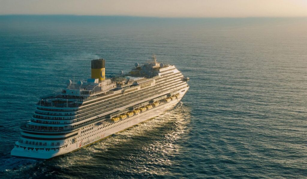 A large Carnival cruise ship sailing through calm ocean waters at sunset, viewed from above.