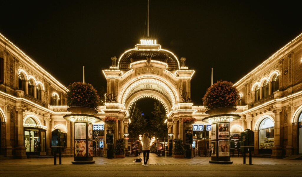 Grand illuminated entrance of Tivoli Gardens in Copenhagen at night, with glowing archways, hanging flower pots, and a person standing in the center holding sparklers.