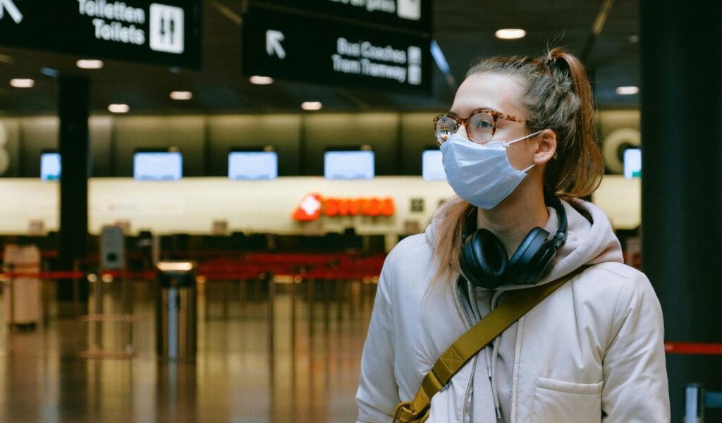 Young woman at airport wearing mask and headphones around neck, ready for travel with music for relaxation.