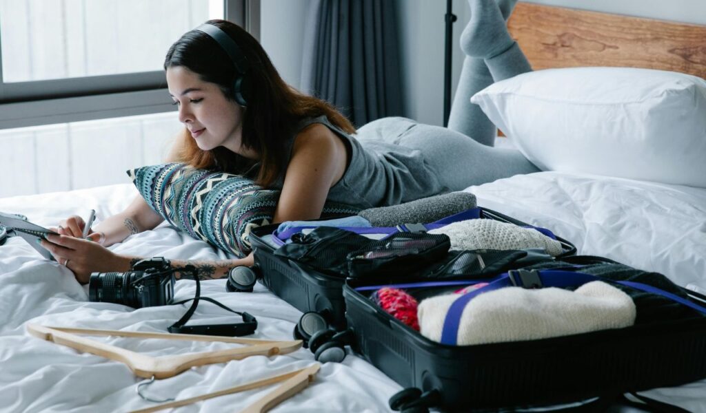 Young woman relaxing on bed with headphones, packing suitcase with travel essentials and personal comfort item