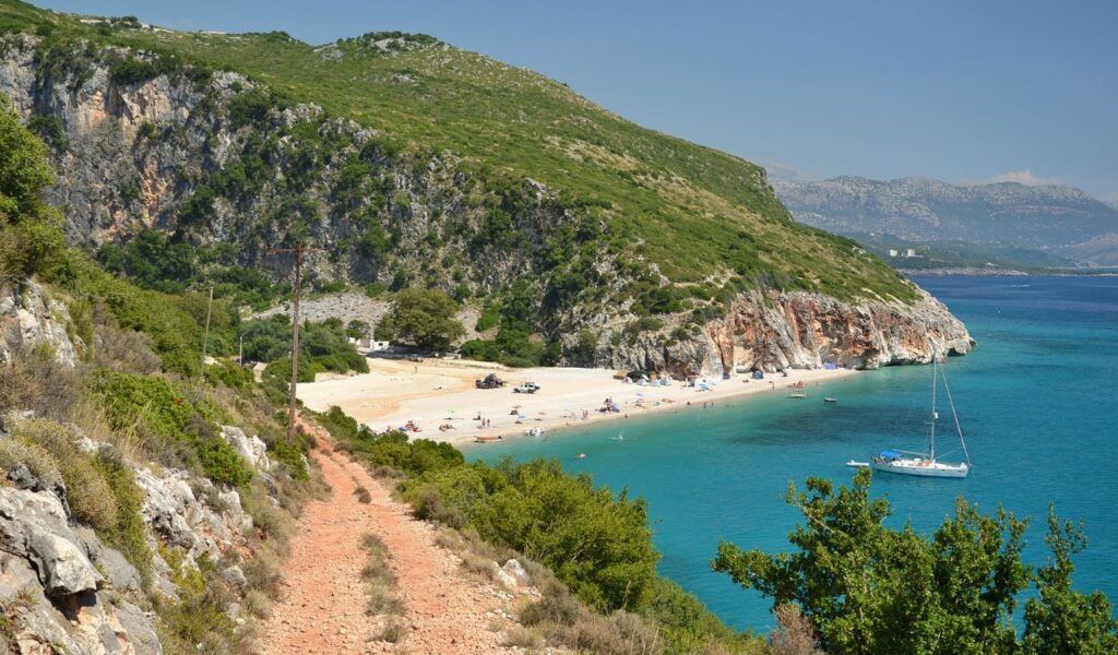 Secluded beach in Croatia surrounded by rocky cliffs, lush greenery, and turquoise waters with people sunbathing and a sailboat anchored nearby.