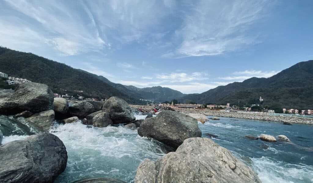 Clear blue waters of a fast-flowing river rushing over large boulders, surrounded by green hills and a small riverside town under a partly cloudy sky.