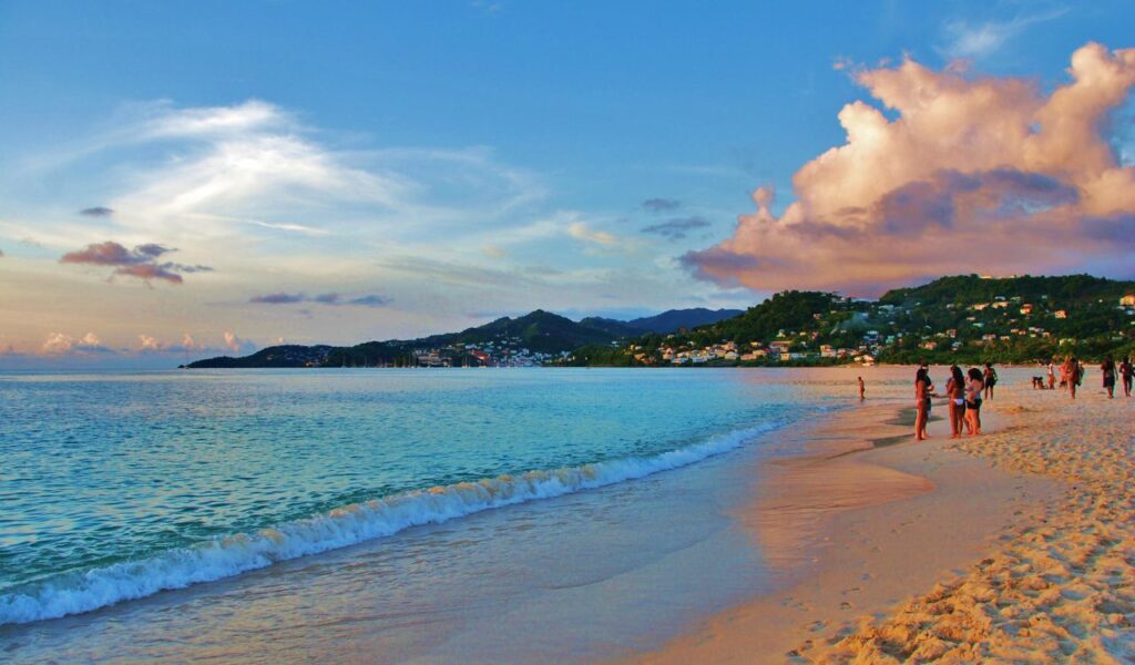 Tropical beach with clear waters, golden sand, and tourists walking along the shoreline under a vibrant sunset sky.