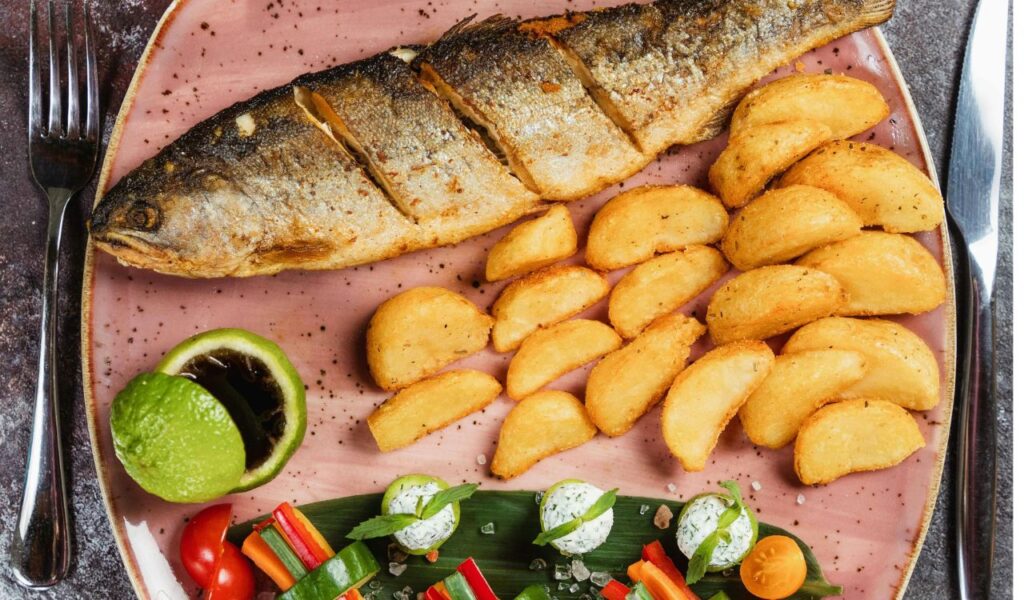 A plate of Culver’s-style fried fish served whole with seasoned potato wedges and colorful vegetable garnishes on a pink ceramic plate.