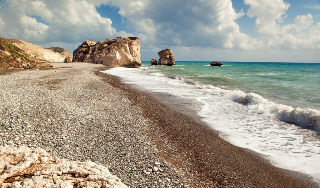 Pebble beach with dramatic rock formations and turquoise waves on the coast of Cyprus under cloudy skies.
