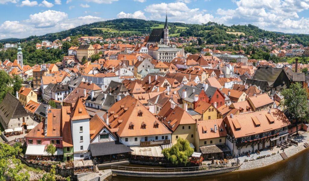 Panoramic view of Český Krumlov’s red-roofed buildings and historic church tower, surrounded by green hills and blue skies.