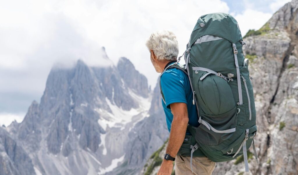 **Alt text:**
Older man with a large hiking backpack stands on a mountain trail, looking out at the dramatic, jagged peaks of the Dolomites partially covered in clouds.