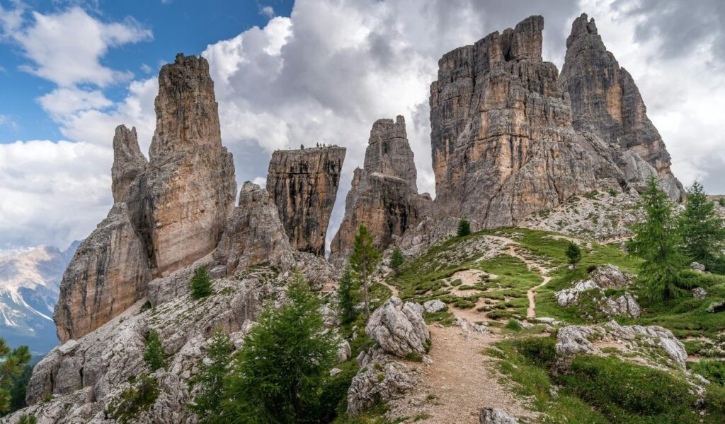 **Alt text:**
Towering stone spires of Cinque Torri rise dramatically from the rocky alpine landscape, surrounded by green shrubs and a winding dirt trail under a partly cloudy sky.